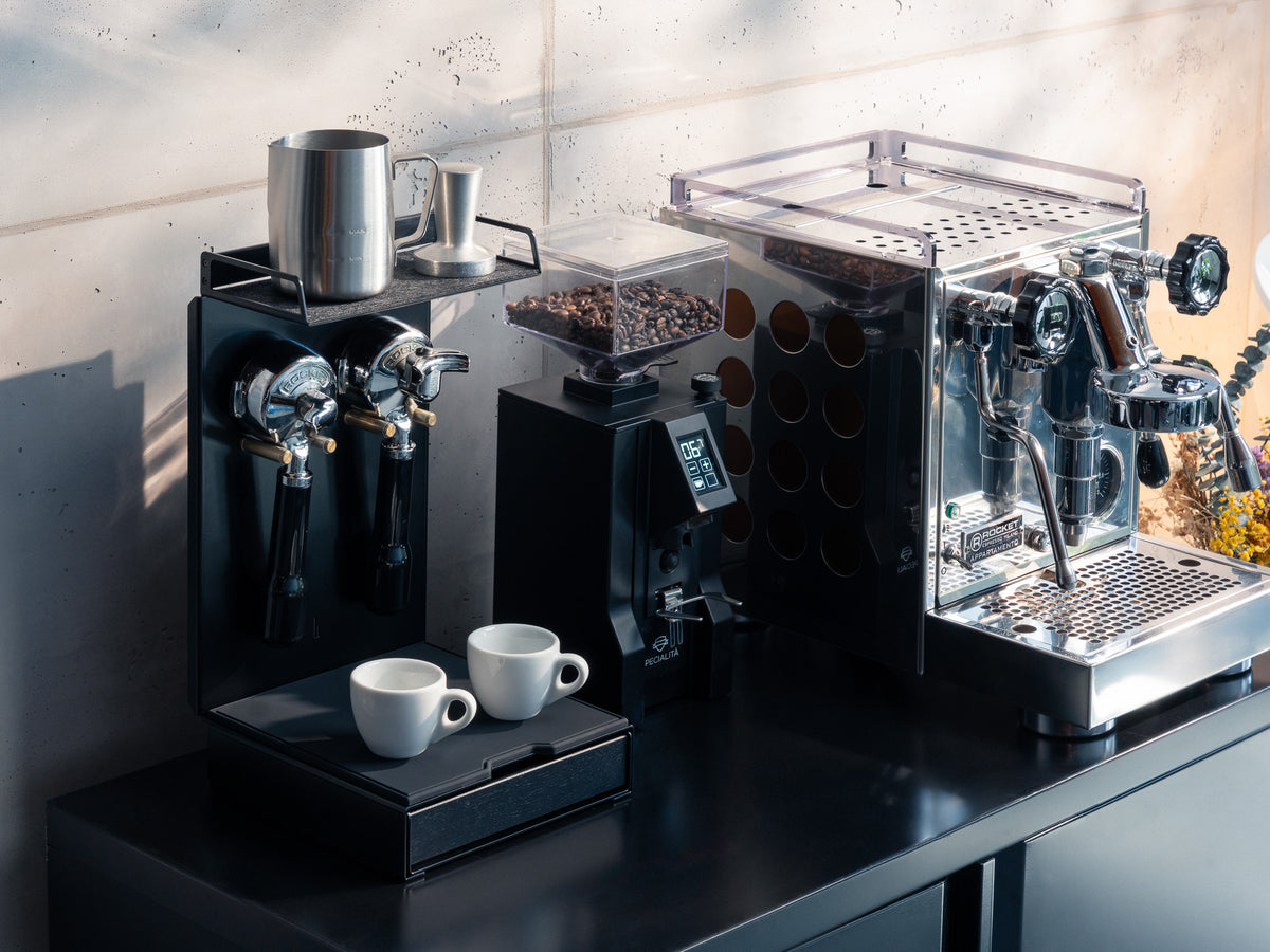 Barista Bar All Black mit Espressomaschine und Mühle auf dunkler Arbeitsplatte vor Betonwand, Sonnenlicht wirft Schatten #material_All Black