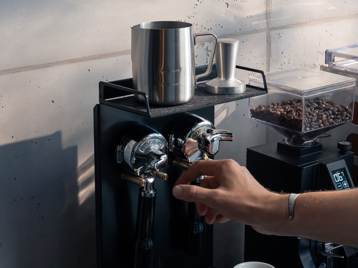 Nahaufnahme: Hand greift nach Siebträger, Milchkännchen und Tamper auf der Ablage, Kaffeebohnen in der Mühle sichtbar @expand #material_All Black