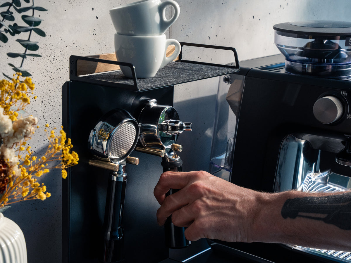Nahaufnahme der Barista Bar Eiche: Hand greift nach Portafilter und Tamper an der Wandhalterung, zwei Tassen auf der Ablage, Trockenblumen im Vordergrund #material_Eiche