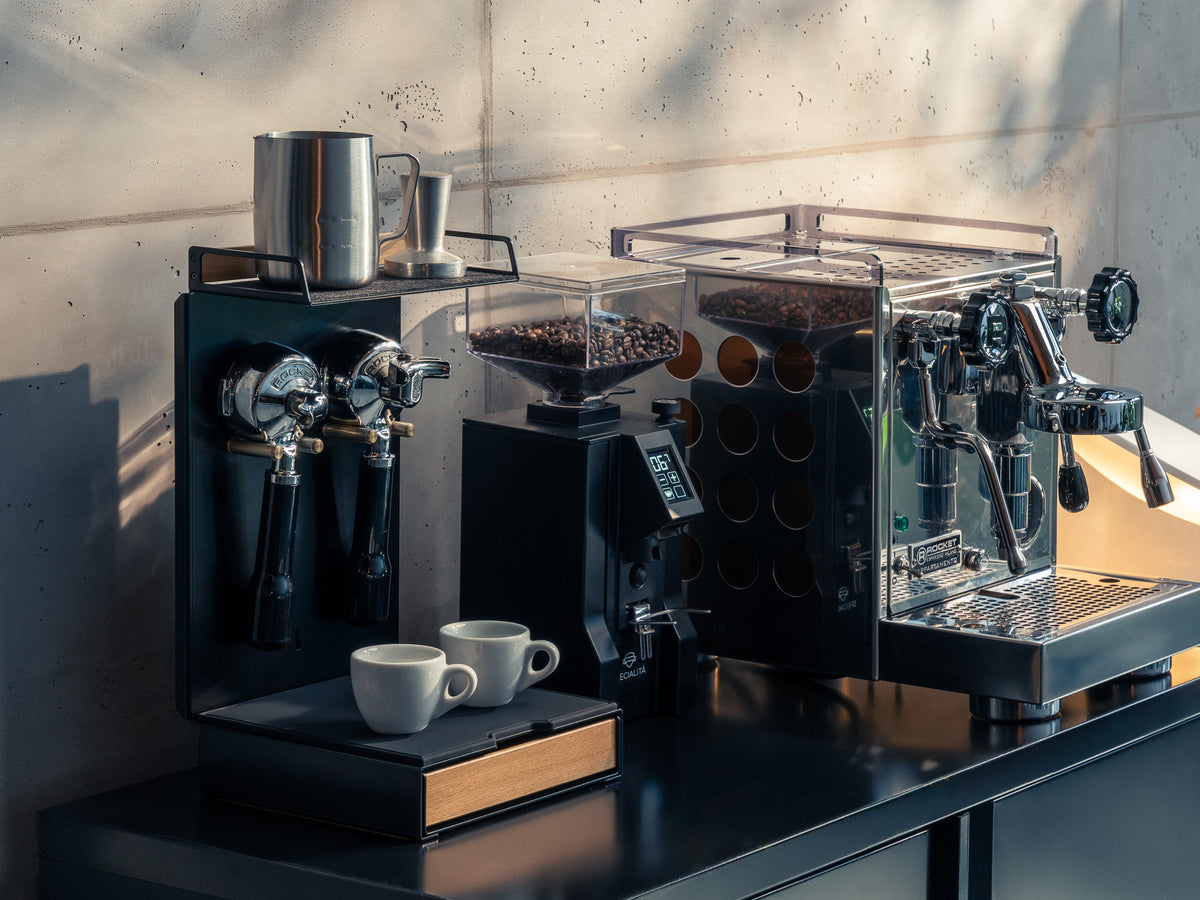 Barista Bar mit Espressomaschine und Mühle auf dunkler Arbeitsplatte vor Betonwand, Sonnenlicht wirft Schatten @expand #material_Eiche
