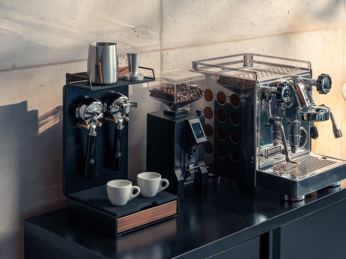 Barista Bar Nussbaum neben Espressomaschine und Kaffeemühle auf dunkler Küchenzeile, zwei Portafilter hängen an der Halterung, zwei Espressotassen auf der ausgezogenen Schublade #material_Amerikanischer Nussbaum