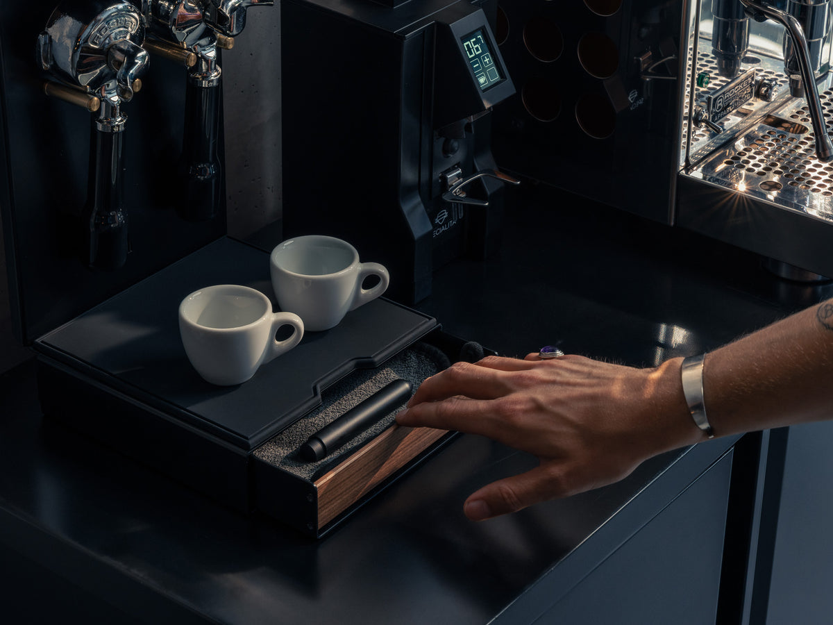 Nahaufnahme: Hand zieht die Schublade der Barista Bar heraus, zwei Espressotassen stehen darauf, im Hintergrund das Display der Kaffeemühle @expand #material_Amerikanischer Nussbaum