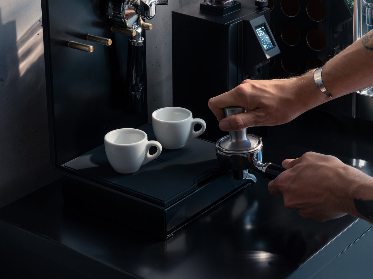 Nahaufnahme: Hände tampern Kaffee im Siebträger auf der Ablagefläche der Barista Bar, zwei Tassen auf der Schublade @expand #material_All Black