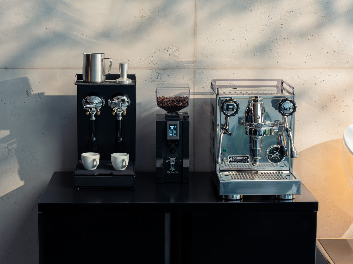 Barista Bar mit Espressomaschine und Mühle auf dunkler Arbeitsplatte vor Betonwand, Sonnenlicht wirft Schatten @expand #material_All Black