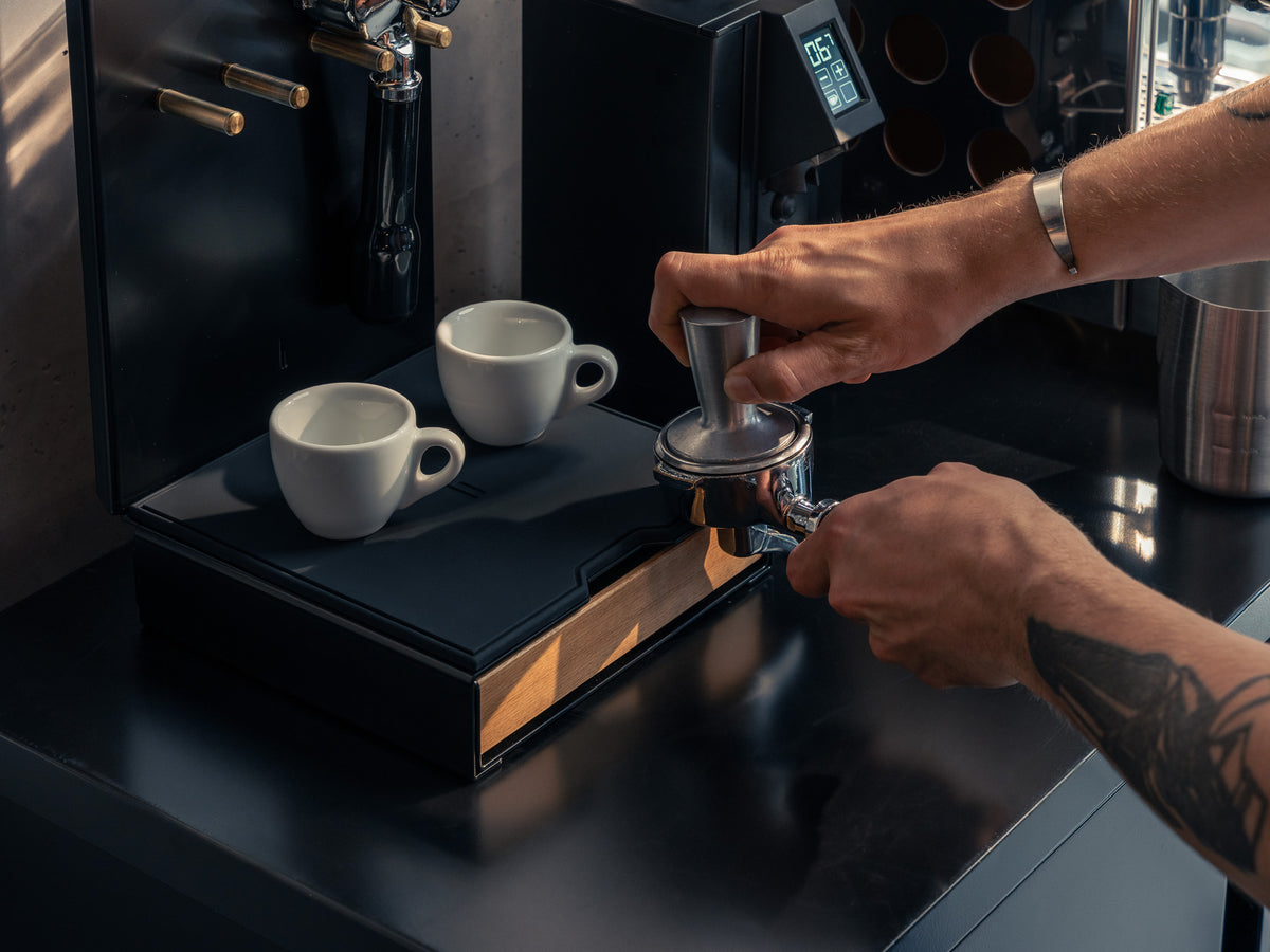 Nahaufnahme: Hände tampern Kaffee, zwei Espressotassen auf der Schublade der Barista Bar, Kaffeemühle mit Bohnenbehälter im Hintergrund @expand #material_Eiche