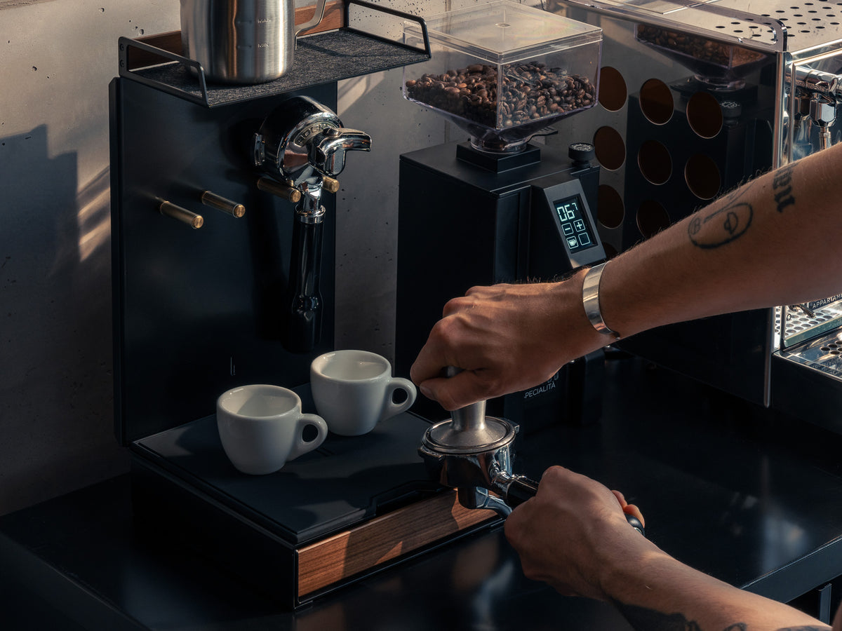 Nahaufnahme: Hände tampern Kaffee im Siebträger auf der Barista Bar, zwei Espressotassen auf der Ablage @expand #material_Amerikanischer Nussbaum