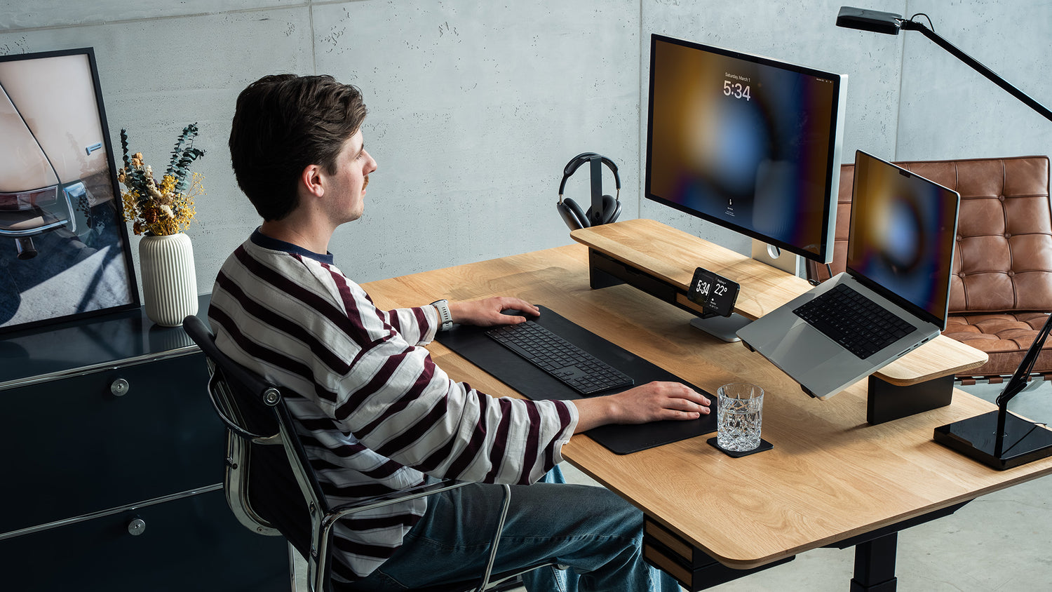 Totalaufnahme eines Office Settings mit BALOLO Setup Cockpit und BALOLO Desk Cockpit. Desk Setup ausgestattet mit Laptop Halter, MagSafe Halterung, Apple Studio Display, Kopfhörerhalter und Desk Pad aus Leder. Person sitzt am Schreibtisch und arbeitet.