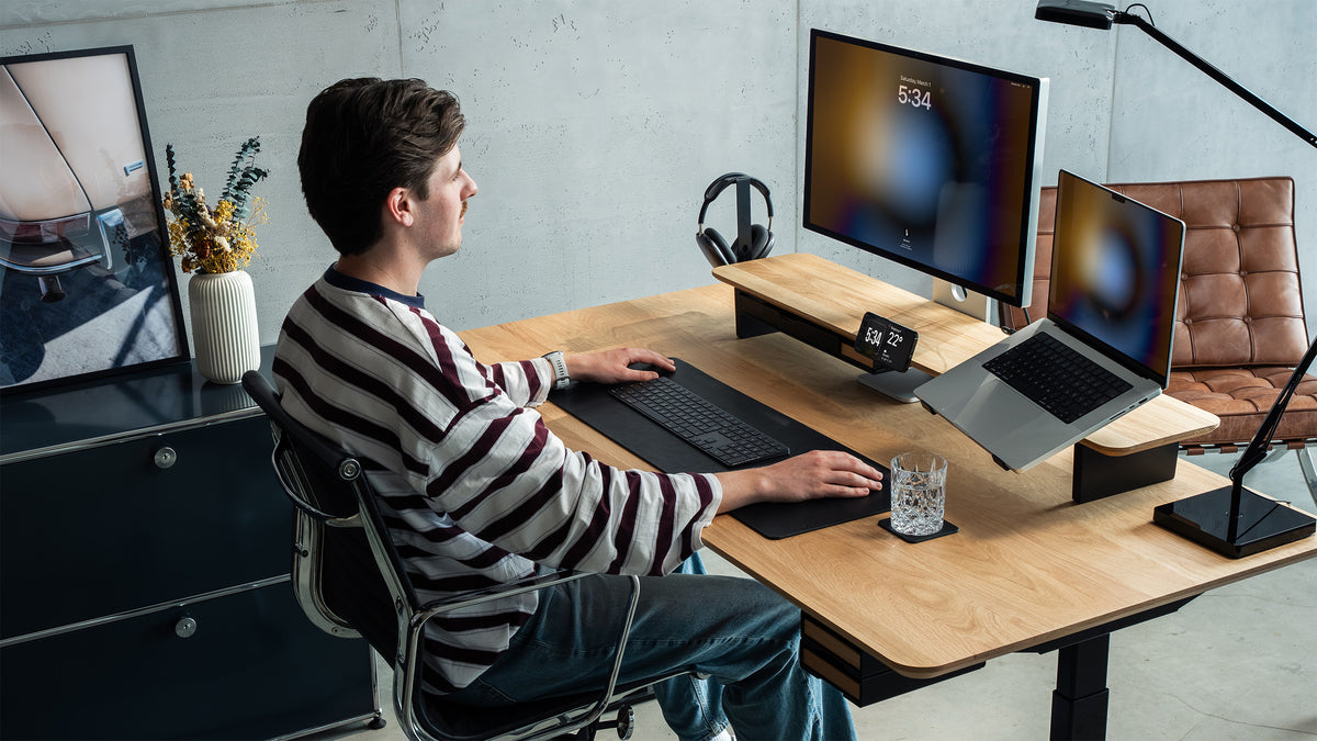 Mann im gestreiften Pullover arbeitet an einem Schreibtisch aus Eiche mit Setup Cockpit Monitorständer, Monitor, Laptop auf Laptophalter, Kopfhörerhalter und schwarzer Leder-Schreibtischunterlage vor einer Betonwand